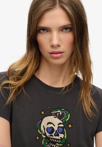Camiseta de color gris oscuro con un diseño frontal de una calavera con ojos azules, adornada con cuentas coloridas, serpientes decorativas y estrellas.