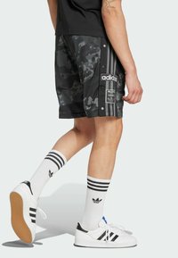 Pantaloni corti neri in camouflage con bottoni a pressione, caratterizzati da tre strisce bianche su ciascun lato; abbinati a calze bianche e sneaker nere e bianche.