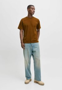 Camiseta de algodón marrón, corte relajado; combinada con jeans de mezclilla azul claro; usando zapatos beige; branding minimalista en el dobladillo de la camiseta.