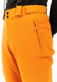 Heldere oranje broek met zwarte knopen, een ritssluiting zijzak en een zwarte tailleband, gedragen met bretels en een zwart bovenstuk.