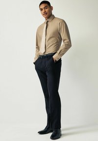 Beige kauluspaita, jossa on terävä kaulus ja pitkät hihat, yhdistettynä kapeisiin tummiin housuihin ja mustiin juhlakengiin. Pinta on sileä kauttaaltaan.