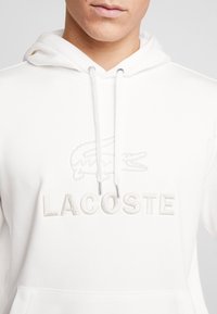 Vit hoodie i mjukt tyg med broderad krokodil-logotyp och "LACOSTE" i matchande tråd. Dragsko och kanguruficka ingår.