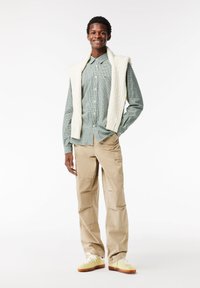 Camicia a quadri verde con pullover in maglia a trecce color crema drappeggiato sopra, abbinata a pantaloni cargo color sabbia e sneakers gialle con suole marroni.