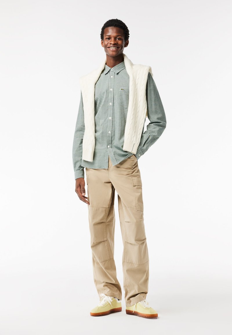 Grün kariertes Hemd mit Knöpfen, über das ein cremefarbener Kabelstrick-Pullover gelegt ist, kombiniert mit beigen Cargo-Hosen und gelben Turnschuhen mit braunen Sohlen.