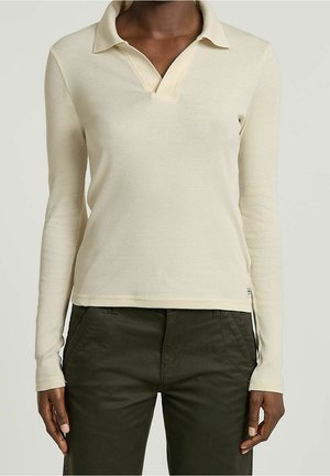 Koszulka polo z długim rękawem w kremowym kolorze o prążkowanej fakturze, z dekoltem w kształcie litery V i kołnierzem. Połączona z ciemnozielonymi spodniami z bocznymi kieszeniami.