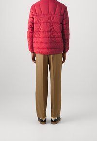 Personne portant une veste matelassée rouge, un pantalon droit marron et des baskets noir et blanc, se tenant devant un fond clair.
