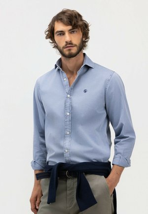 Hombre con cabello rizado y barba, vistiendo camisa azul claro abotonada, pantalones beige y suéter oscuro atado alrededor de la cintura, de pie con la mano en el bolsillo.