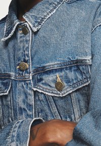 Jeansjacka i ljusblått med texturerad yta, med två bröstfickor, metallknappar och fransdetaljer längs kragen och ärmsluten.