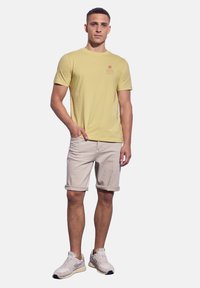 Světle žluté tričko s krátkým rukávem a grafickým potiskem vpředu, spárované s béžovými krátkými kalhotami a šedými teniskami, stojící v neutrální póze.