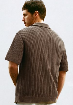 Rövid sötét hajú férfi barna bordázott rövid ujjú pulóvert visel, oldalról néz egy egyszínű világos háttér előtt.