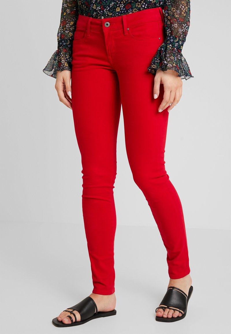 Pantalones vaqueros ajustados rojos con una textura suave, diseño de cinco bolsillos y un botón plateado en la cinturilla, combinados con sandalias negras tipo slides.