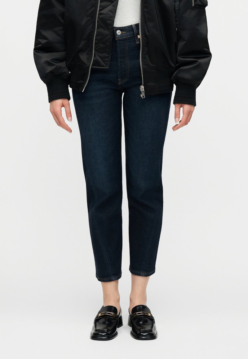 Άτομο που φοράει σκούρο μπλε τζιν, μαύρα μοκασίνια, λευκή μπλούζα και μαύρο μπουφάν bomber, όρθιο μπροστά σε απλό φόντο.