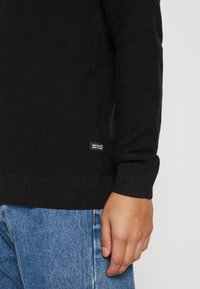 Pull en maille noire à texture côtelée, avec un col rond et une petite étiquette logo sur le côté. Un jean en denim bleu est partiellement visible.