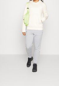 Persona che indossa una felpa bianca, pantaloni della tuta grigio chiaro, sneakers nere e porta una piccola borsa a tracolla verde chiaro su uno sfondo semplice.