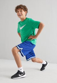 Camiseta verde de Nike con logo blanco, pantalones cortos azules con acentos blancos, zapatillas negras y calcetines blancos. Posicionado en una zancada dinámica.