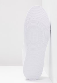 Semelle d'une basket blanche avec des motifs circulaires texturés et un logo embossé discret au centre, photographiée sur des surfaces blanches.