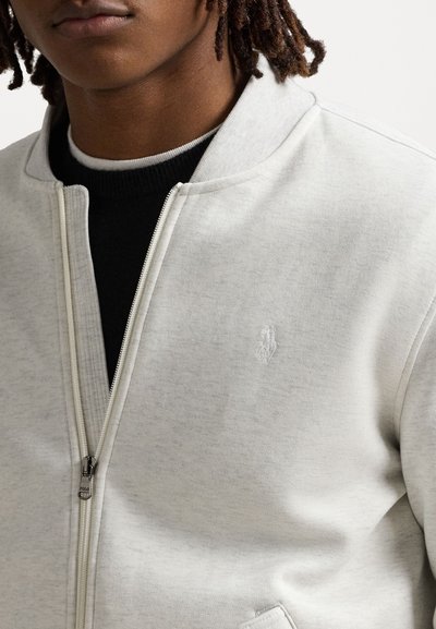 Svetlošedá bundu na zips s hladkým povrchom, rebrovaným golierom a vyšitým logom na hrudi. Obsahuje zips a bočné vrecká.