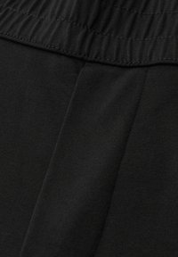 Nærbillede af sorte stofbukser med elastisk taljebånd og detaljeret midtersøm.