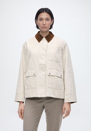 Nő krémszínű dzsekiben, barna gallérral és három első zsebbel, barna-fehér kockás nadrággal, egyszerű háttér előtt állva.