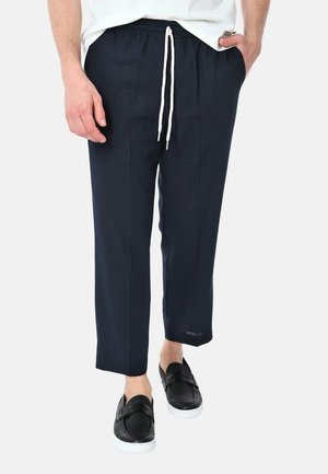 Uomo che indossa pantaloni blu navy con coulisse bianca e scarpe slip-on nere, in piedi con una mano in tasca e camicia bianca.