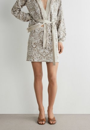 Femme portant une robe beige et blanche à motif cachemire avec des manches longues et une ceinture en tissu, associée à des talons ouverts marron.