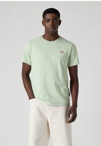 Ανοιχτοπράσινο βαμβακερό t-shirt με στρογγυλή λαιμόκοψη, κοντά μανίκια και μικρό κόκκινο λογότυπο Levi's στο αριστερό στήθος. Το μοντέλο το συνδυάζει με παντελόνι σε εκρού απόχρωση.