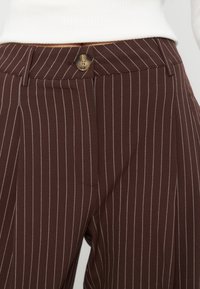 Pantalon marron avec fines rayures blanches verticales, fermeture à boutons devant, passants de ceinture, et partie d'un haut blanc visible au-dessus de la taille.