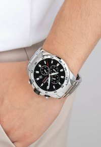 Montre en acier inoxydable argenté avec un cadran noir, des index lumineux et trois sous-cadrans. Dispose d'un guichet de date et d'un bracelet en acier inoxydable.