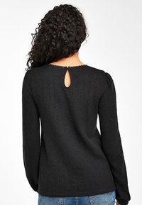 Haut noir texturé à manches longues, avec une découpe en forme de trou de serrure à l'arrière et une bordure en dentelle autour du col. Coupe décontractée.