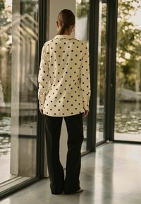 Chemise à manches longues couleur crème avec un motif à pois noirs. Associée à un pantalon large noir. L'arrière-plan présente de grandes fenêtres en verre et un décor naturel.