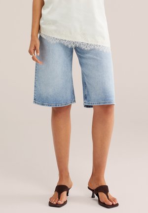 Žena v svetlomodrých rifle kraťasoch po kolená, bielom top s čipkovanými lemami a hnedých sandáloch na podpätku stojí na jednofarebnom pozadí.