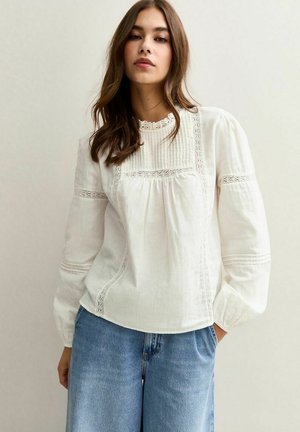Blusa de lino blanca con mangas largas abullonadas, detalles plisados y acentos de encaje en el cuello y las costuras, combinada con jeans de color azul claro.