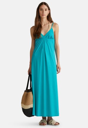 A long, sleeveless teal dress with a deep V-neck and gathered waist, paired with a textured shoulder bag and light-colored sandals.