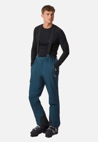 Pantaloni da sci blu con bretelle nere regolabili, tasche laterali, rinforzi alle ginocchia e una texture morbida. Gli accessori includono stivali neri.