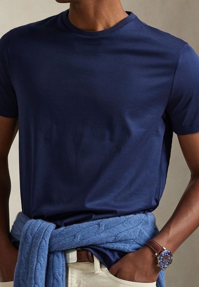 Άνδρας που φορά μπλε ναυτικό μπλουζάκι, ανοιχτό μπεζ παντελόνι με καφέ ζώνη, μπλε πλεκτό πουλόβερ δεμένο γύρω από τη μέση και ρολόι χειρός με μπλε καντράν και καφέ λουρί.