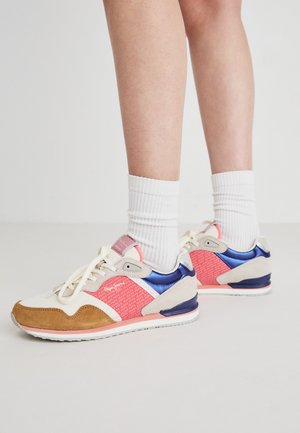 Adidași sport cu un superior crem, accente din plasă roz, vârf din suede maro, accente strălucitoare albastre și șireturi albe, purtați cu șosete albe.