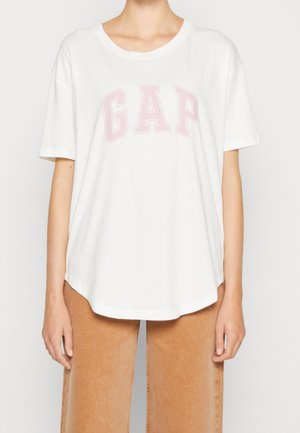Camiseta blanca de manga corta hecha de algodón con cuello redondo. Presenta el logo "GAP" en un suave rosa claro en el pecho. Textura suave.