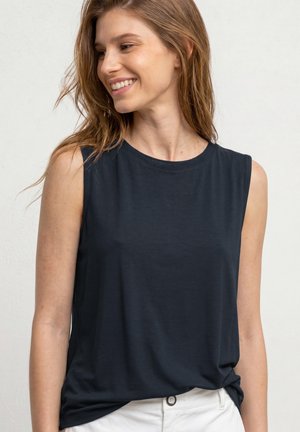 Femme souriante aux longs cheveux portant un haut noir sans manches rentré dans un pantalon blanc, debout devant un fond clair uni.