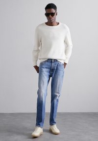 Fehér bordás kötött pulóver, világoskék, megfakult farmernadrág és krém színű sportcipő. A modell napszemüveget visel, háttérben egy egyszínű háttérrel.