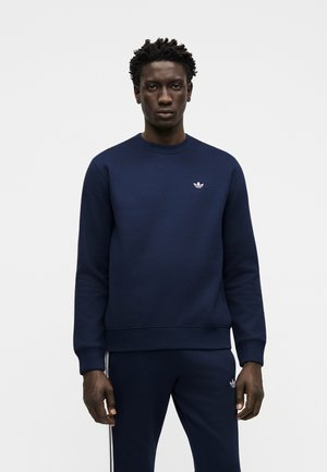 Mand iført navyblå Adidas sweatshirt og matchende bukser med hvide trefoil-logoer, stående mod en ensfarvet lys baggrund.