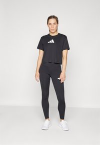 Svart avkortet t-skjorte med hvit logo på brystet, kombinert med svarte leggings med en liten hvit logo. Modellen har på seg hvite treningessko.