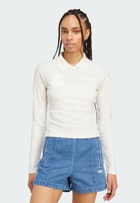 Long-sleeve white polo with collar, featuring an embroidered logo, paired with blue denim shorts, displaying a casual sporty style.