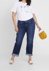 Valkoinen t-paita, jossa on musta kukkapainatus ja teksti, yhdistetty tummansiniset farkut, joiden lahkeet on käännetty, sekä pieni keltainen käsilaukku.