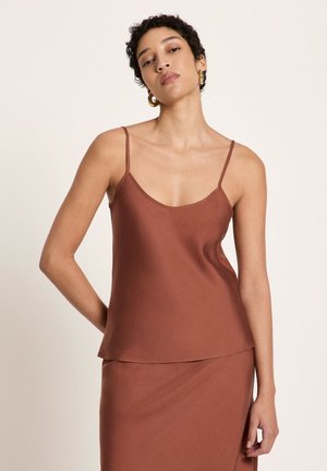 Frau mit kurzen lockigen Haaren, die ein braunes ärmelloses Oberteil und einen passenden Rock trägt, vor einem schlichten hellen Hintergrund stehend.