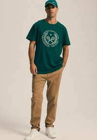 Zielona bawełniana koszulka z logo stowarzyszenia tenisowego, beżowe spodnie i białe sneakersy. Model nosi dopasowaną zieloną czapkę. Prosty design.