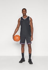 Juodas krepšinio marškinėliai su baltais kraštais, derinami su pilkais šortais, turinčiais spalvotus dryžius. Žaidėjas laiko krepšinio kamuolį ir yra apsirengęs juodais batais.