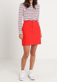 Jupe en coton rouge avec fermeture à boutons sur le devant, associée à un haut à rayures à manches longues. Des baskets blanches complètent le look avec un style décontracté.