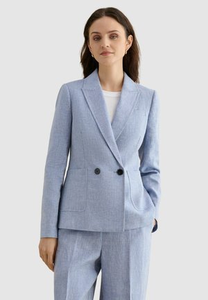 Frau trägt einen hellblauen, maßgeschneiderten Blazer und passende Hose, mit einem weißen Top darunter, steht vor einem schlichten Hintergrund.