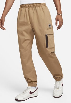 Herren beige Cargohosen mit elastischem Bund und Bündchen, seitlicher Reißverschlusstasche, kombiniert mit weißen und dunkelroten Nike Sneakern.