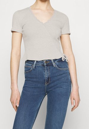 Frau trägt ein hellgraues Wickeloberteil mit kurzen Ärmeln und wellenförmigen Kanten sowie hoch taillierte blaue Jeans.